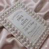 An ivory silk save the date card embroidered with faux pearls and rhinestones, with details printed with black foil on a pearl shimmery cardstock.