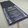 A modern wedding invitation made of clear acrylic, resting on a burlap surface. The invitation features elegant white screen-printed text and is framed by white floral scrollwork borders. A black folio is visible in the blurred background.