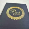 A top-down view of an open, square black silk invitation box. The interior lid features a matching horizontal silk pocket holding a Acrylic card with the names in elegant black script. The base of the box is lined with plush, gathered black fabric. The box is set against a textured white fabric background.