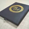 A top-down view of an open, square black silk invitation box. The interior lid features a matching horizontal silk pocket holding a Acrylic card with the names in elegant black script. The base of the box is lined with plush, gathered black fabric. The box is set against a textured white fabric background.