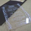 A modern wedding invitation made of clear acrylic, resting on a burlap surface. The invitation features elegant white screen-printed text and is framed by white floral scrollwork borders. A black folio is visible in the blurred background.