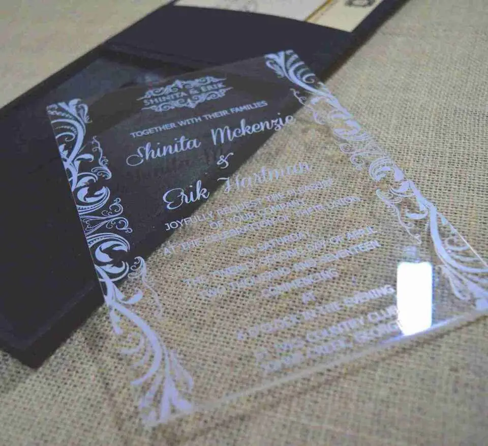 A modern wedding invitation made of clear acrylic, resting on a burlap surface. The invitation features elegant white screen-printed text and is framed by white floral scrollwork borders. A black folio is visible in the blurred background.