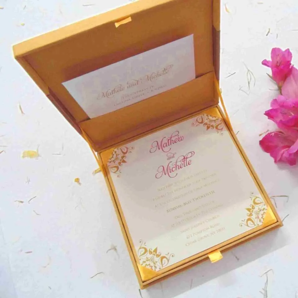 A top-down view of an open gold silk invitation box. The interior lid features a matching silk pocket holding a white card with the names "Mathew and Michelle" in elegant gold script. The base of the box contains a formal invitation card with gold-foil floral accents in the corners. The box is set against a white background and is decorated with scattered dried pink flower petals.