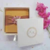 A top-down view of a square gold silk invitation box resting on a white surface. The lid is wrapped with a wide, bronze-toned satin ribbon, featuring a large, ornate oval brooch at the center encrusted with pink and clear crystals. The scene is decorated with scattered dried petals and bright pink gladiolus flowers along the right side.