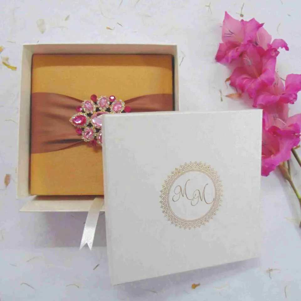 A top-down view of a square gold silk invitation box resting on a white surface. The lid is wrapped with a wide, bronze-toned satin ribbon, featuring a large, ornate oval brooch at the center encrusted with pink and clear crystals. The scene is decorated with scattered dried petals and bright pink gladiolus flowers along the right side.
