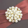 A high-angle shot of a rectangular, dark brown silk invitation box resting on a light-colored marbled surface. A wide, burgundy satin ribbon is wrapped horizontally around the center, gathered in the middle by a large, circular brooch. The brooch features a cluster of creamy white pearls interspersed with small, gold-toned floral accents set with tiny crystals. A small ribbon pull-tab is visible at the bottom edge of the box.