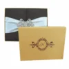 A top-down, centered shot of a chocolate brown silk gatefold invitation box. A wide, pleated white satin ribbon wraps horizontally around the middle, secured at the center by a large, ornate butterfly brooch encrusted with shimmering clear crystals. The background is solid white.