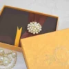 A high-angle shot of a rectangular, dark brown silk invitation box resting on a light-colored marbled surface. A wide, burgundy satin ribbon is wrapped horizontally around the center, gathered in the middle by a large, circular brooch. The brooch features a cluster of creamy white pearls interspersed with small, gold-toned floral accents set with tiny crystals. A small ribbon pull-tab is visible at the bottom edge of the box.