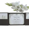 An open, dark brown folio wedding invitation set against a white background, accented by a branch of white gladiolus flowers at the top. The central panel holds a white invitation card with elegant black script and ornate corner flourishes, announcing the marriage of Victoria Marie and Patric Johnson. The side panels feature pockets containing RSVP and detail cards with matching decorative motifs.