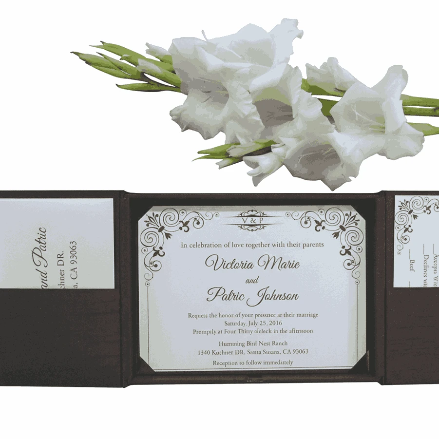 An open, dark brown folio wedding invitation set against a white background, accented by a branch of white gladiolus flowers at the top. The central panel holds a white invitation card with elegant black script and ornate corner flourishes, announcing the marriage of Victoria Marie and Patric Johnson. The side panels feature pockets containing RSVP and detail cards with matching decorative motifs.