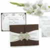 An open, dark brown folio wedding invitation set against a white background, accented by a branch of white gladiolus flowers at the top. The central panel holds a white invitation card with elegant black script and ornate corner flourishes, announcing the marriage of Victoria Marie and Patric Johnson. The side panels feature pockets containing RSVP and detail cards with matching decorative motifs.