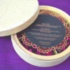 An open, round ivory-colored favor box resting on a textured purple surface. The box is divided into four compartments, each filled with vibrant magenta dried flower petals. Next to the box lies its lid, covered in an elegant lace floral pattern, and a circular black menu card with a decorative gold laser-cut border.