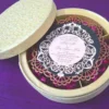 An open, round ivory-colored favor box resting on a textured purple surface. The box is divided into four compartments, each filled with vibrant magenta dried flower petals. Next to the box lies its lid, covered in an elegant lace floral pattern, and a circular black menu card with a decorative gold laser-cut border.