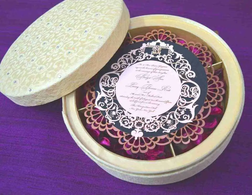 An open, round ivory-colored favor box resting on a textured purple surface. The box is divided into four compartments, each filled with vibrant magenta dried flower petals. Next to the box lies its lid, covered in an elegant lace floral pattern, and a circular black menu card with a decorative gold laser-cut border.