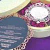 An open, round ivory-colored favor box resting on a textured purple surface. The box is divided into four compartments, each filled with vibrant magenta dried flower petals. Next to the box lies its lid, covered in an elegant lace floral pattern, and a circular black menu card with a decorative gold laser-cut border.