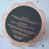 A top-down view of a circular wedding menu card resting on a light, textured surface. The card is matte black with elegant rose-gold or copper text detailing courses for Salad, Entrée, and Dessert. The menu is framed by an intricate, laser-cut copper border with a delicate scalloped lace pattern.