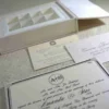 A flat-lay of a white wedding gift suite. It features a closed box with a crystal brooch, an open box base containing nine gold-wrapped chocolate truffles in a grid, a formal invitation for Mathew and Michelle, an RSVP card, and an envelope, all accented by pink flowers.