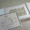 A flat-lay of a white wedding gift suite. It features a closed box with a crystal brooch, an open box base containing nine gold-wrapped chocolate truffles in a grid, a formal invitation for Mathew and Michelle, an RSVP card, and an envelope, all accented by pink flowers.