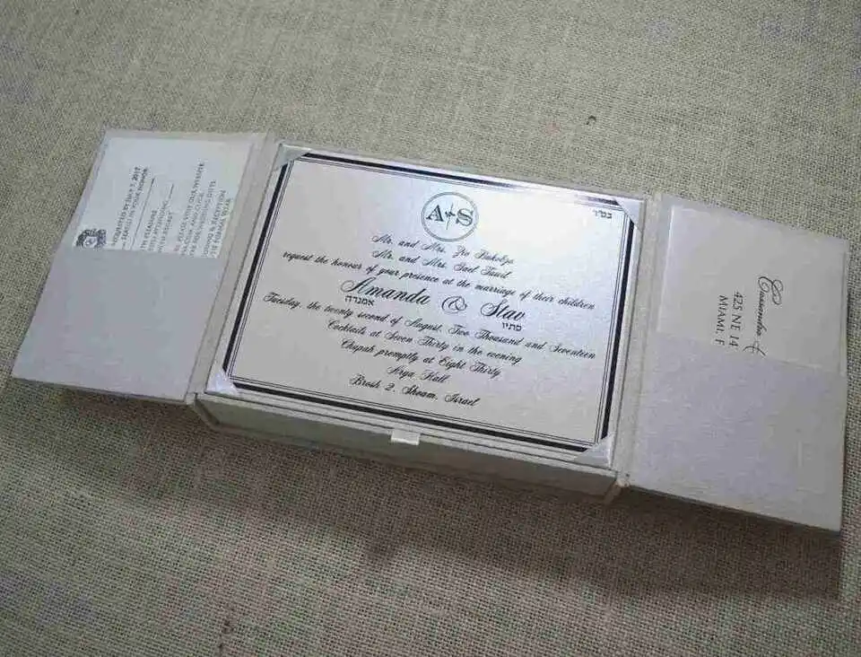 A top-down view of an open, champagne-colored silk invitation box. The interior lid features a matching horizontal silk pocket holding a white card printed with the name "Mathew and Michelle" in elegant gold script. The base of the box contains a formal wedding invitation card with gold-foil floral accents in the corners. A stem of vibrant pink gladiolus flowers lies to the right of the box on a textured white surface.