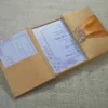 An open, gold silk-textured gatefold wedding invitation box resting on a burlap surface. The central panel displays the wedding invitation for "Lada Yana," featuring elegant cursive text and a delicate floral illustration in the corner. The left flap contains a pocket holding a white RSVP card with lace-like detailing. On the right, a shimmering orange satin ribbon is visible, secured by an ornate, crystal-encrusted silver brooch. The image is taken from a low side angle, highlighting the depth and texture of the luxury invitation suite.
