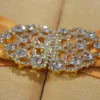A rectangular, gold silk gatefold wedding invitation box shown from a perspective angle. A wide, shimmering gold satin ribbon wraps horizontally around the box, gathered at the center by an ornate, oval-shaped silver brooch encrusted with clear, sparkling crystals. The box rests on a neutral-toned, textured burlap or linen surface. The lighting emphasizes the fine, horizontal weave of the silk and the warm, metallic tones of the invitation suite.