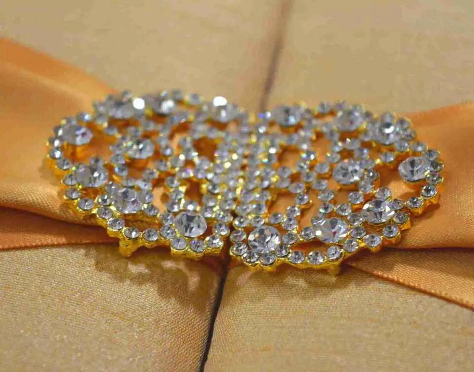 A rectangular, gold silk gatefold wedding invitation box shown from a perspective angle. A wide, shimmering gold satin ribbon wraps horizontally around the box, gathered at the center by an ornate, oval-shaped silver brooch encrusted with clear, sparkling crystals. The box rests on a neutral-toned, textured burlap or linen surface. The lighting emphasizes the fine, horizontal weave of the silk and the warm, metallic tones of the invitation suite.