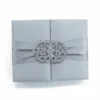 A top-down, centered shot of a square, silver silk gatefold invitation box. A matching silver satin ribbon wraps horizontally around the center, gathered by a large, ornate filigree brooch encrusted with shimmering clear crystals. The entire box sits against a clean, solid white background.