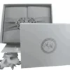 A top-down, centered shot of a square, silver silk gatefold invitation box. A matching silver satin ribbon wraps horizontally around the center, gathered by a large, ornate filigree brooch encrusted with shimmering clear crystals. The entire box sits against a clean, solid white background.