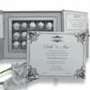 An open, light silver wedding invitation box set against a white background. The central compartment contains a grid of twelve chocolates wrapped in silver foil. A white invitation card, featuring elegant black script and ornate corner flourishes for "Farah & Amir," leans against the front of the box. The right side flap of the box contains an RSVP card with a matching design. A soft white flower is visible in the bottom left corner.