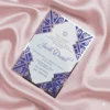 A sophisticated Bar Mitzvah invitation for presented on a shimmering, draped blush pink satin background. The white card features navy blue text and a at the top. It is encased in a **polished silver acrylic frame** featuring intricate Art Deco-style laser-cut designs in the corners, which reveal a **royal blue** backing. The overall design combines classic religious elements with a modern, architectural aesthetic.