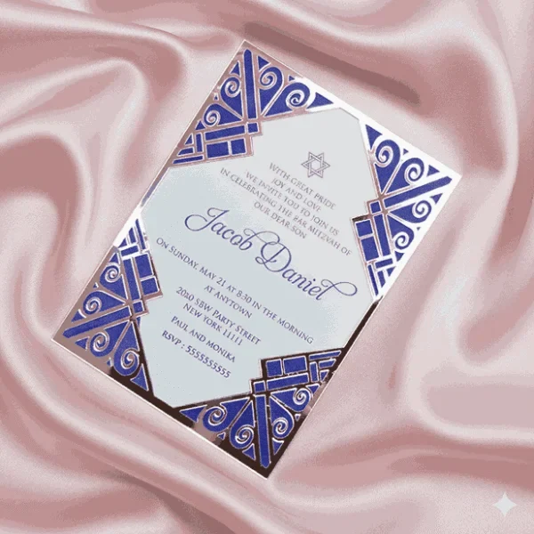A sophisticated Bar Mitzvah invitation for presented on a shimmering, draped blush pink satin background. The white card features navy blue text and a at the top. It is encased in a **polished silver acrylic frame** featuring intricate Art Deco-style laser-cut designs in the corners, which reveal a **royal blue** backing. The overall design combines classic religious elements with a modern, architectural aesthetic.