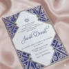 A sophisticated Bar Mitzvah invitation for presented on a shimmering, draped blush pink satin background. The white card features navy blue text and a at the top. It is encased in a **polished silver acrylic frame** featuring intricate Art Deco-style laser-cut designs in the corners, which reveal a **royal blue** backing. The overall design combines classic religious elements with a modern, architectural aesthetic.
