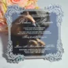 A square wedding invitation made of a highly reflective silver mirror acrylic material. The invitation features an ornate, white baroque-style floral border in each of the four corners and along the scalloped edges The mirror surface is so reflective that the photographer holding a camera is visible in the center of the card. A bouquet of peach-colored roses is partially visible in the blurred background on the left.