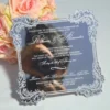A square wedding invitation made of a highly reflective silver mirror acrylic material. The invitation features an ornate, white baroque-style floral border in each of the four corners and along the scalloped edges The mirror surface is so reflective that the photographer holding a camera is visible in the center of the card. A bouquet of peach-colored roses is partially visible in the blurred background on the left.