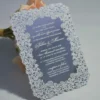 This image features a high-end silver mirror acrylic wedding invitation with an intricate white laser-cut lace border. The card is shown at an angle, leaning against a bouquet of soft peach roses on a white surface. The invitation features elegant white printed text with the details.