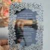 This image features a high-end silver mirror acrylic wedding invitation with an intricate white laser-cut lace border. The card is shown at an angle, leaning against a bouquet of soft peach roses on a white surface. The invitation features elegant white printed text with the details.