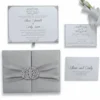 A comprehensive wedding invitation suite in silver and white, displayed against a solid white background. * **Bottom Left:** A closed, square silver silk gatefold box with a pleated silver satin ribbon and a large, ornate crystal-encrusted filigree brooch. * **Top Left:** The main invitation card for "Oliver Edwards & Emily Luna" featuring elegant black cursive and a decorative crest. * **Top Right:** A coordinating RSVP card with matching scrollwork. * **Bottom Right:** A detail card with the couple's names and the venue address in Cleveland, Ohio. * **Accents:** A small portion of a white flower is visible in the upper right corner.