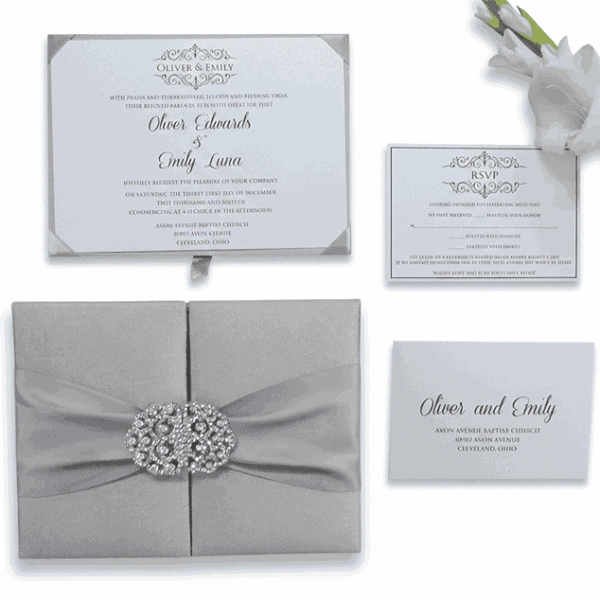 A comprehensive wedding invitation suite in silver and white, displayed against a solid white background. * **Bottom Left:** A closed, square silver silk gatefold box with a pleated silver satin ribbon and a large, ornate crystal-encrusted filigree brooch. * **Top Left:** The main invitation card for "Oliver Edwards & Emily Luna" featuring elegant black cursive and a decorative crest. * **Top Right:** A coordinating RSVP card with matching scrollwork. * **Bottom Right:** A detail card with the couple's names and the venue address in Cleveland, Ohio. * **Accents:** A small portion of a white flower is visible in the upper right corner.
