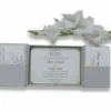 An open, silver-grey gatefold wedding invitation box displayed against a white background. The center compartment holds a white invitation card for "Oliver Edwards & Emily Luna," featuring elegant cursive text and a decorative crest at the top. The two side panels have pockets containing coordinating RSVP and detail cards. A branch of soft white gladiolus flowers lies horizontally across the top, framing the invitation suite.