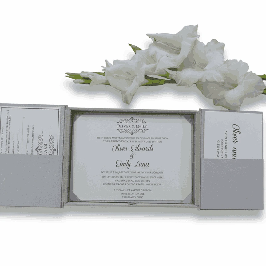 An open, silver-grey gatefold wedding invitation box displayed against a white background. The center compartment holds a white invitation card for "Oliver Edwards & Emily Luna," featuring elegant cursive text and a decorative crest at the top. The two side panels have pockets containing coordinating RSVP and detail cards. A branch of soft white gladiolus flowers lies horizontally across the top, framing the invitation suite.