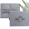 A top-down shot of a closed, square silver silk gatefold invitation box on a white background. A matching silver satin ribbon is cinched at the center by a large, ornate crystal-encrusted filigree brooch. To the right, a vertical stem of white gladiolus flowers with green buds adds a delicate, natural accent.
