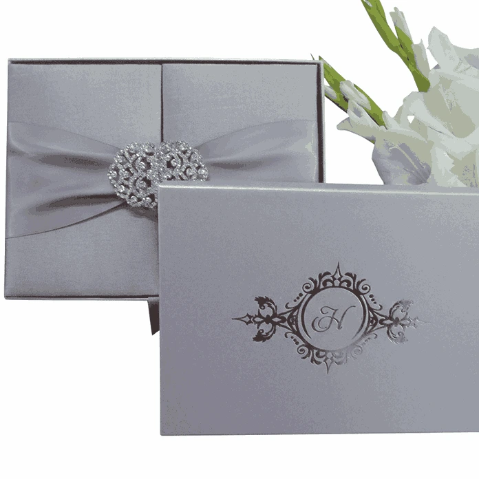 A top-down shot of a closed, square silver silk gatefold invitation box on a white background. A matching silver satin ribbon is cinched at the center by a large, ornate crystal-encrusted filigree brooch. To the right, a vertical stem of white gladiolus flowers with green buds adds a delicate, natural accent.