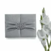 A top-down shot of a closed, square silver silk gatefold invitation box on a white background. A matching silver satin ribbon is cinched at the center by a large, ornate crystal-encrusted filigree brooch. To the right, a vertical stem of white gladiolus flowers with green buds adds a delicate, natural accent.
