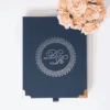 A top-down, centered shot of a navy blue textured invitation box on a white surface. The lid features a silver-foiled monogram with the initials "D" and "R" enclosed in a circular, lace-like ornate border. The bottom corners of the box are protected by polished rose gold metal caps, and a small navy pull-tab is visible at the bottom edge. In the top right corner, a bouquet of peach-colored roses adds a soft, elegant accent.