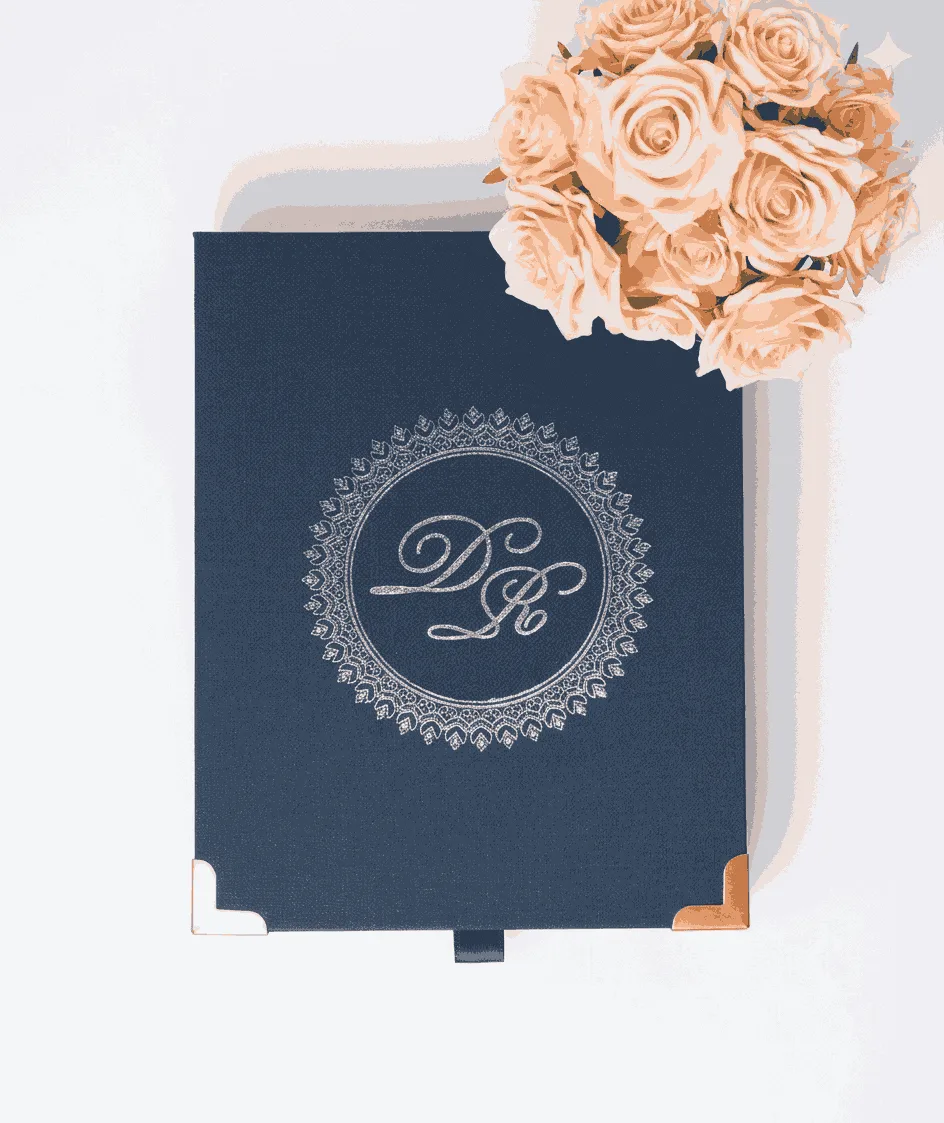 A top-down, centered shot of a navy blue textured invitation box on a white surface. The lid features a silver-foiled monogram with the initials "D" and "R" enclosed in a circular, lace-like ornate border. The bottom corners of the box are protected by polished rose gold metal caps, and a small navy pull-tab is visible at the bottom edge. In the top right corner, a bouquet of peach-colored roses adds a soft, elegant accent.