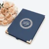A top-down, centered shot of a navy blue textured invitation box on a white surface. The lid features a silver-foiled monogram with the initials "D" and "R" enclosed in a circular, lace-like ornate border. The bottom corners of the box are protected by polished rose gold metal caps, and a small navy pull-tab is visible at the bottom edge. In the top right corner, a bouquet of peach-colored roses adds a soft, elegant accent.