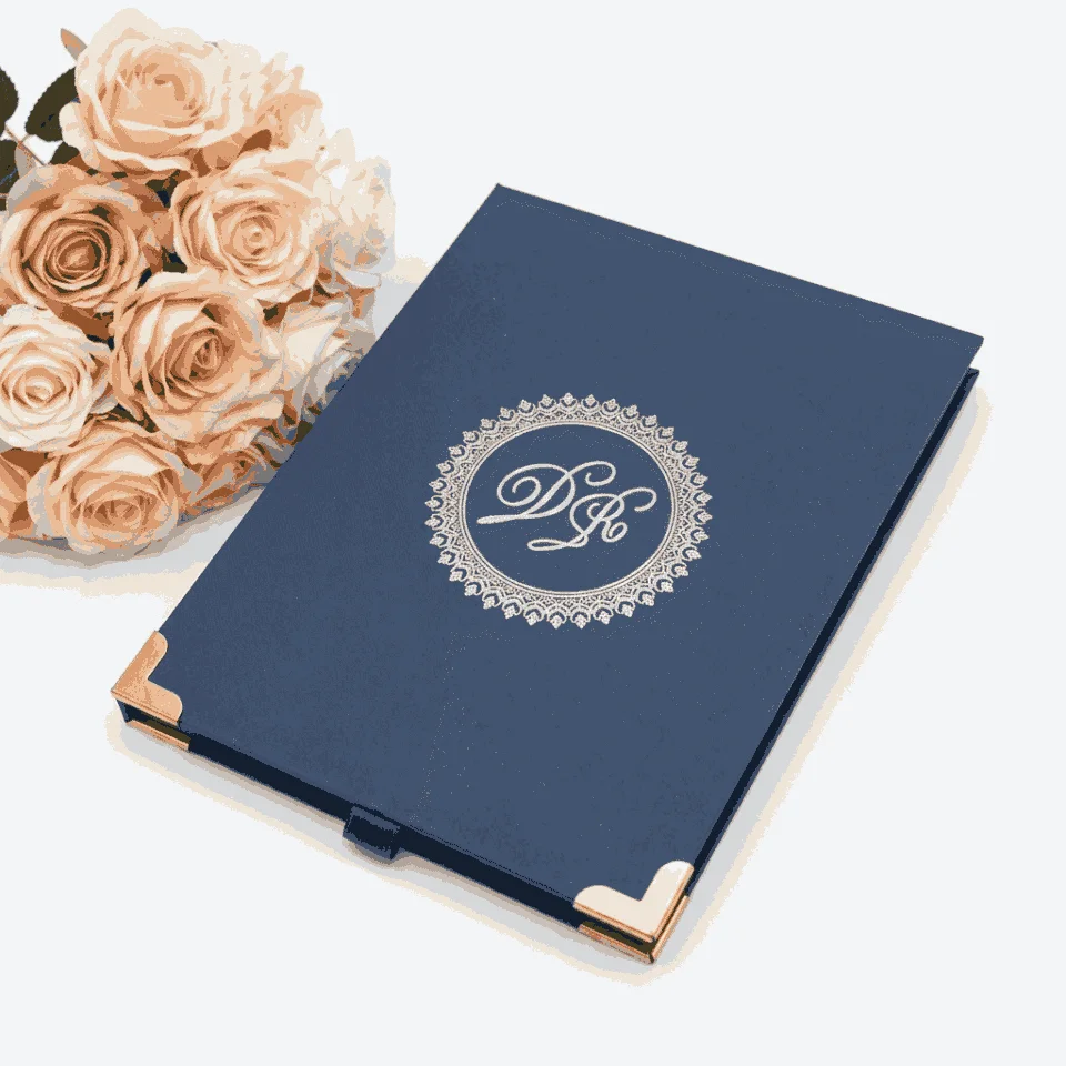 A top-down, centered shot of a navy blue textured invitation box on a white surface. The lid features a silver-foiled monogram with the initials "D" and "R" enclosed in a circular, lace-like ornate border. The bottom corners of the box are protected by polished rose gold metal caps, and a small navy pull-tab is visible at the bottom edge. In the top right corner, a bouquet of peach-colored roses adds a soft, elegant accent.