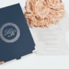 An overhead, angled shot of a wedding invitation suite on a white surface. To the left is a navy blue textured invitation box featuring a silver-foiled "DR" monogram inside an ornate circular border and rose gold metal corner accents. To the right, a clear acrylic invitation card rests partially over a bouquet of peach roses. The acrylic card is engraved with white text for the wedding of "Jacqueline & Anthony," detailing an event at the Verona Event Center. The elegant design includes delicate white flourishes in the corners of the clear card.