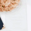 A high-angle, vertical shot of a clear acrylic wedding invitation resting on a white surface. The invitation is engraved with elegant white script and block lettering for the wedding of "Jacqueline & Anthony," featuring delicate floral border flourishes in the corners. The top left of the card overlaps a lush bouquet of peach-colored roses. In the bottom left corner, the edge of a navy blue invitation box with a polished gold corner cap is visible.