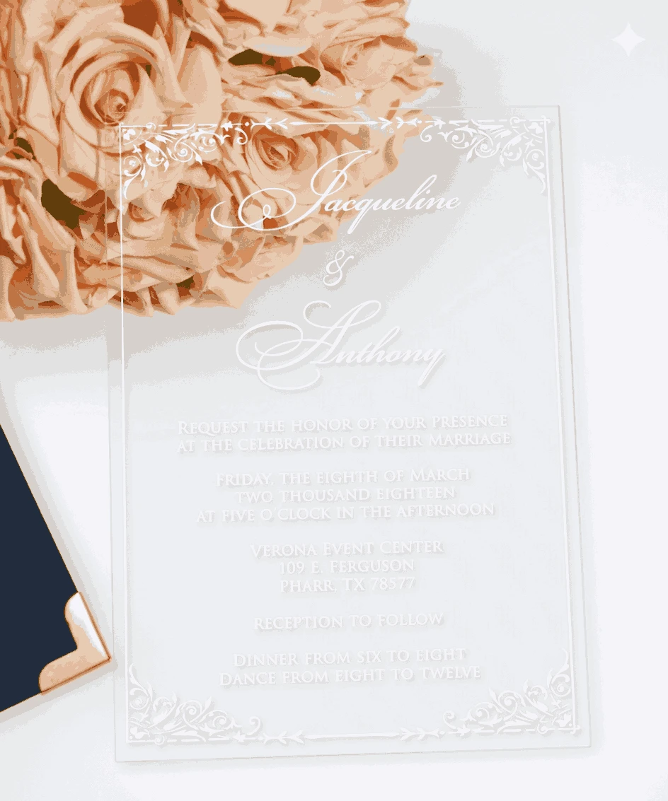 A high-angle, vertical shot of a clear acrylic wedding invitation resting on a white surface. The invitation is engraved with elegant white script and block lettering for the wedding of "Jacqueline & Anthony," featuring delicate floral border flourishes in the corners. The top left of the card overlaps a lush bouquet of peach-colored roses. In the bottom left corner, the edge of a navy blue invitation box with a polished gold corner cap is visible.